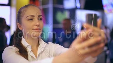 一个年轻的女人坐在快餐店里，拍自拍，在智能手机屏幕上看她的照片。